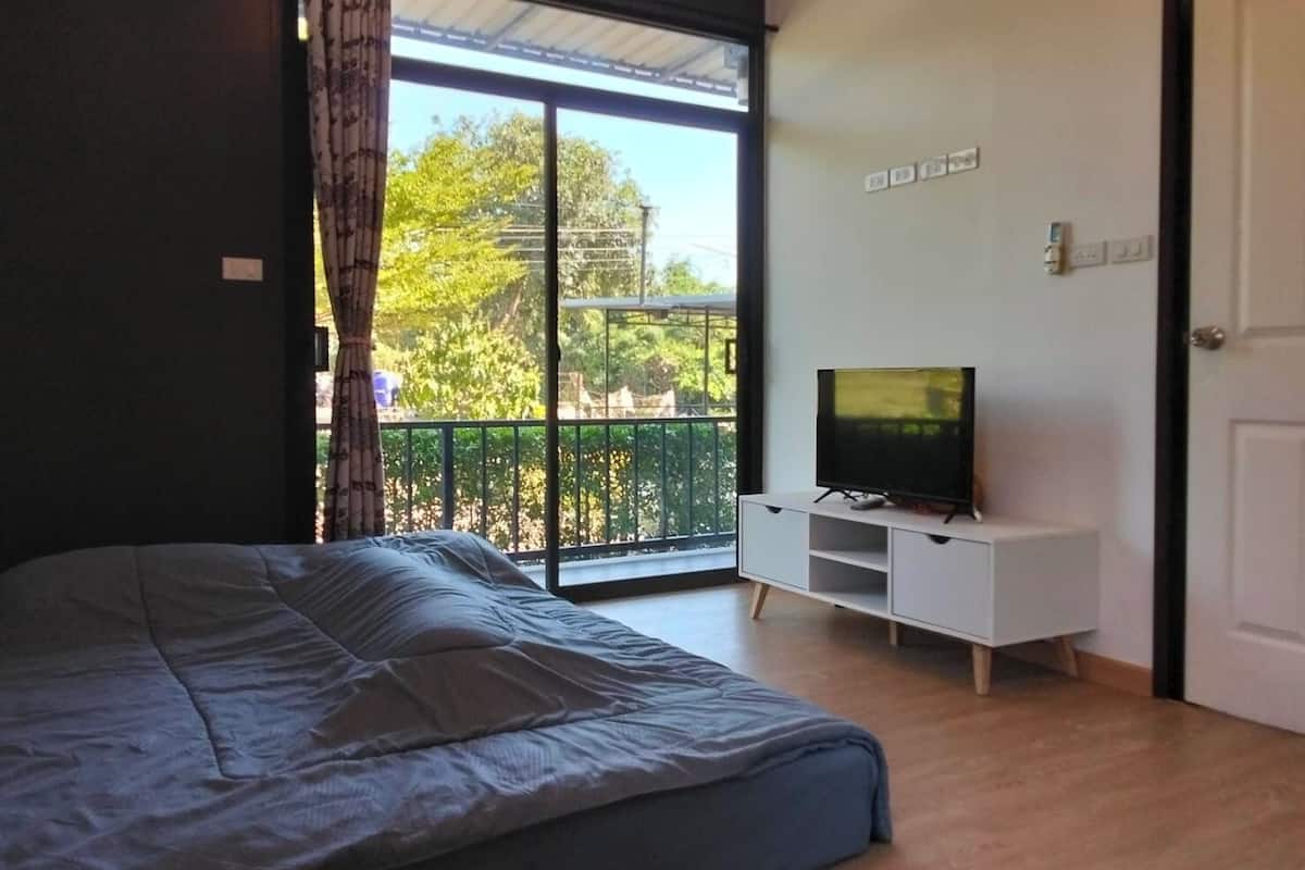 A bedroom with a bed, a TV on a white cabinet, and a balcony with a view of trees and a fence.