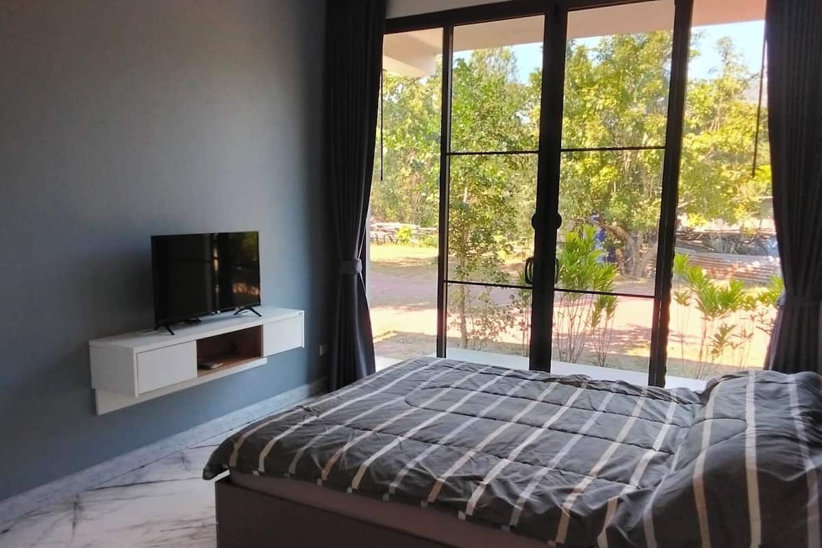 A bedroom with a bed, a TV on a white shelf, and a large window with a view of greenery.