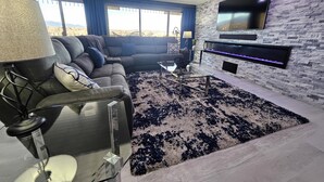 A modern living room with a sectional sofa, a glass coffee table, a flat-screen TV mounted on a stone wall, and a large window with a view of the desert.