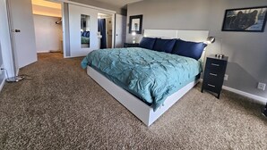 A bedroom with a bed, a nightstand, a lamp, a dresser, and a door leading to another room.
