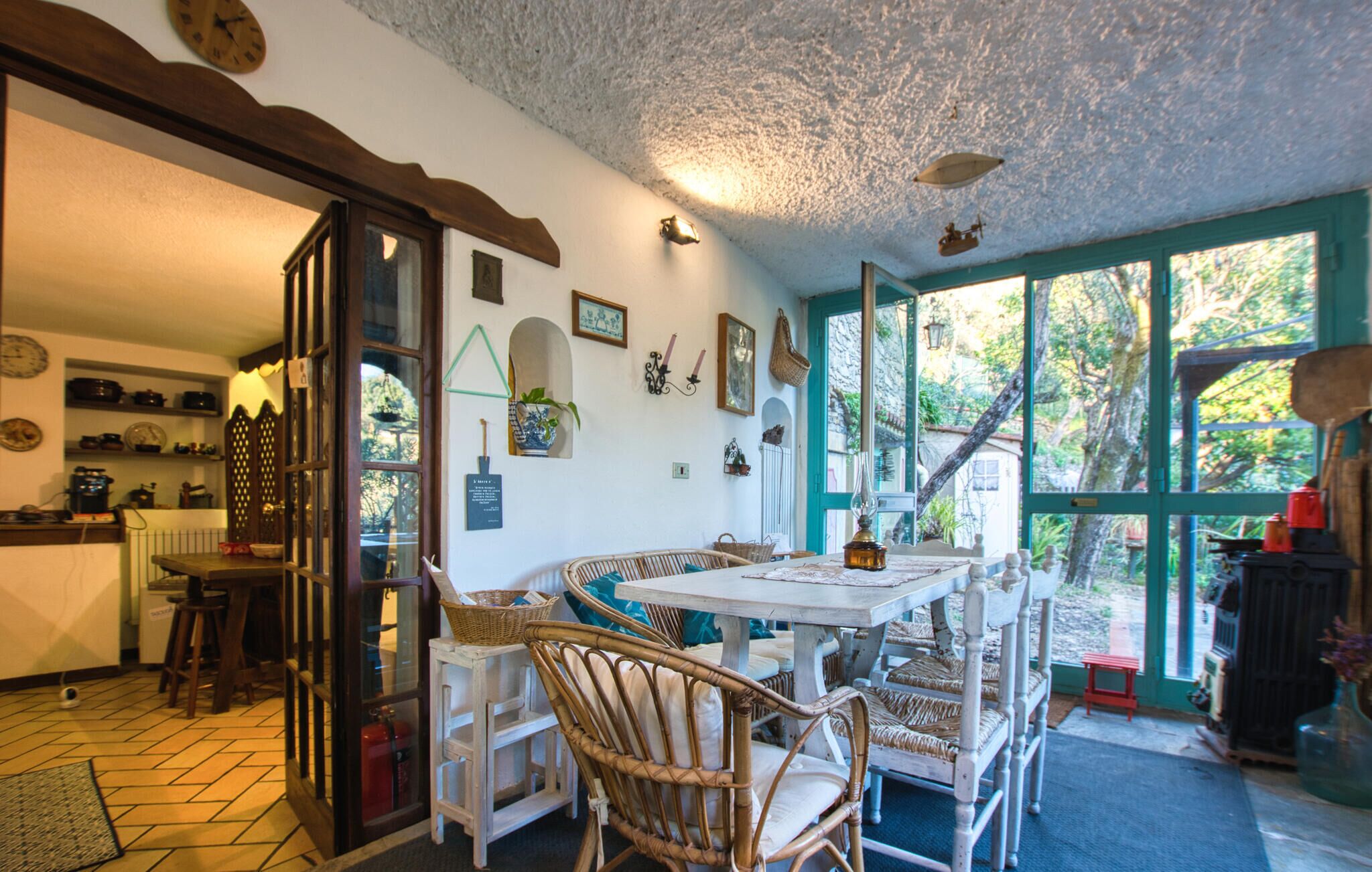 A cozy dining area with wicker chairs and a white table, a kitchen with shelves of dishes, and a large window with a view of greenery.