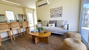 A modern living space with a sofa, coffee table, and wicker chairs. A kitchen area with a refrigerator and a dining table is visible.