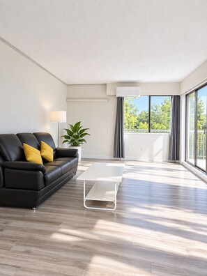 A modern living room with a black leather sofa, a white coffee table, a potted plant, and a view of greenery through large windows. - Modern 1 Bedroom Suite with Parking (North Vancouver)