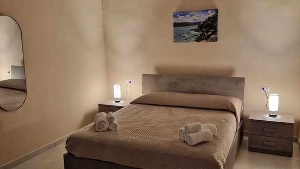 Un dormitorio moderno con una cama grande, dos mesitas de noche con lámparas, un espejo en la pared y un cuadro enmarcado de un paisaje.