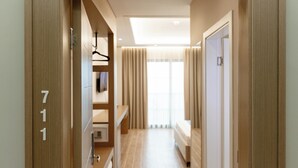 A hotel hallway with wooden flooring, a ceiling light, a bed, and a window with curtains.