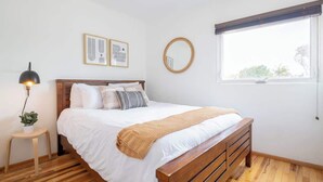 A bedroom with a wooden bed, white bedding, a small bedside table with a lamp, and a window with a view of greenery. - Boho Beauty! Super Central | Steps to Beach & Bay (San Diego)