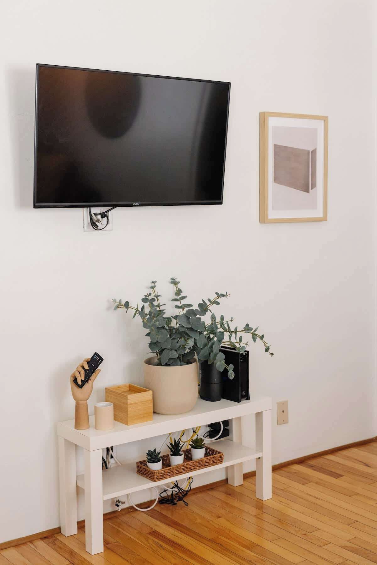 A flat-screen TV mounted on a white wall, a framed picture to the right, and a white console table below with a potted plant, a wooden sculpture, and a remote control.