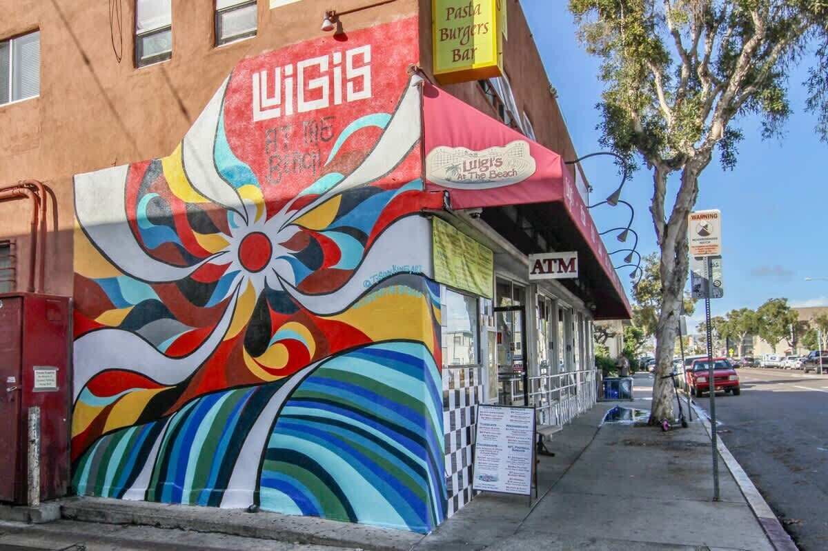 Une fresque colorée orne le mur d’un bâtiment, sur lequel on peut voir une enseigne indiquant « Luigi’s At The Beach » et un distributeur automatique de billets.