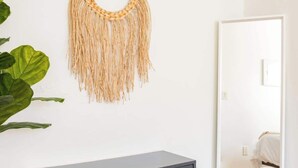 Una camera moderna con un settimino in legno scuro, un arazzo tessuto e un grande specchio che riflette un letto e una porta.