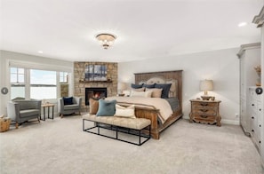 A bedroom with a stone fireplace, a large bed with multiple pillows, a bench, a nightstand, and a dresser. - Unplug in Style at This Premier Minnesota Lakefront Haven (Buffalo)