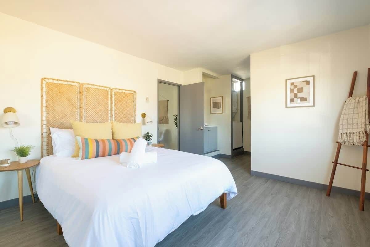 Ein Hotelzimmer mit einem großen Bett, einem Nachttisch mit Lampe, einer kleinen Pflanze und einem Bild an der Wand.
