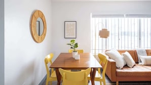 Een eetgedeelte met een houten tafel en gele stoelen, een bruine bank met kussens, een potplant en een ingelijst kunstwerk aan de muur.