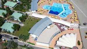 Eine Vogelperspektive zeigt ein Resort mit mehreren Schwimmbecken, einer großen überdachten Fläche und zahlreichen Strandstühlen.