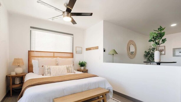 Un dormitorio con cama, ventilador de techo, mesita de noche, lámpara, banco, ventana con persianas, espejo y planta.