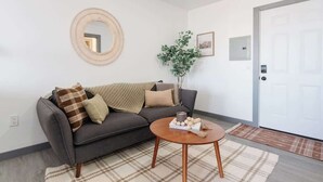 Una sala de estar con un sofá gris, una mesa redonda de centro, un espejo, una planta y una puerta.