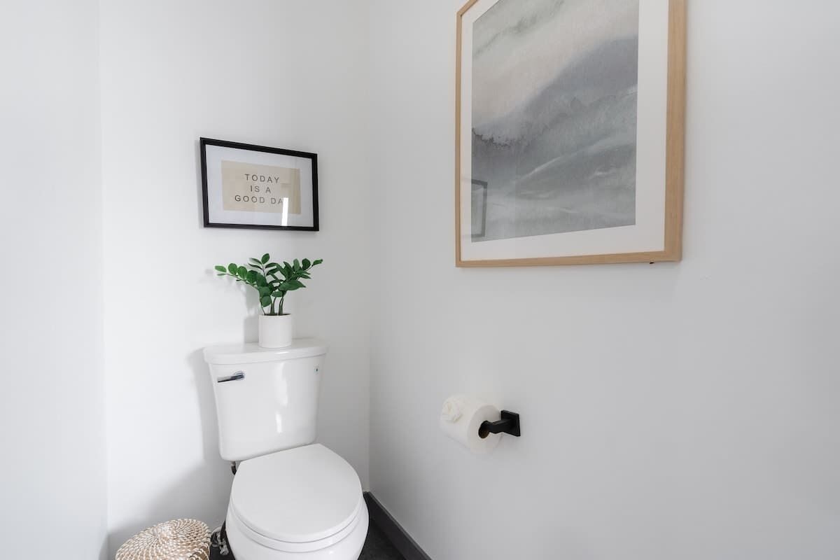 A modern bathroom with a white toilet, a framed inspirational quote, a potted plant, and a framed abstract painting.