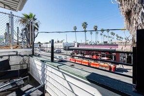 A view from a balcony overlooking a street with palm trees, a parking lot, and a building with a red awning. - Stylish & Comfy near Beach & Pier ~ Balcony ~ Pkg! (San Diego)