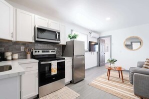 A modern kitchen with white cabinets, a stainless steel refrigerator, a microwave, and an oven. There is a small wooden table with a plant, and a gray sofa in the living area. - Stylish & Comfy near Beach & Pier ~ Balcony ~ Pkg! (San Diego)