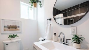 Een moderne badkamer met een wit wastafel, een ronde spiegel en een toilet. Er staan twee potplanten, hangt een lijst met een foto aan de muur, en een raam zorgt voor natuurlijk licht.