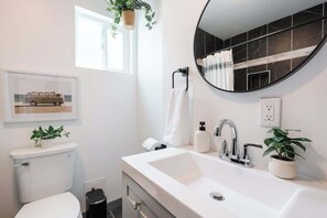 A modern bathroom with a white sink, a round mirror, and a toilet. There are two potted plants, a framed picture on the wall, and a window providing natural light. - Super Central Retreat | Steps to Everything! (San Diego)