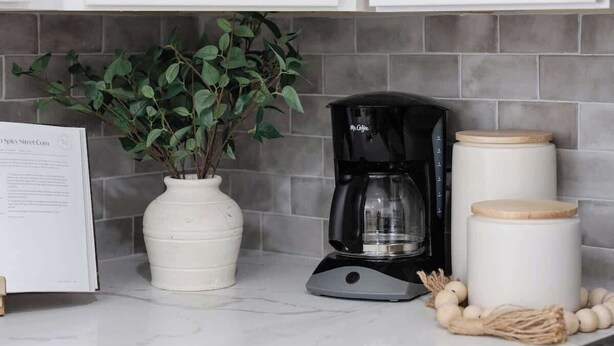 Een keuken met witte kastjes, een zwarte koffiezetapparaat, een witte vaas met groene bladeren, en een bordje op het aanrecht.