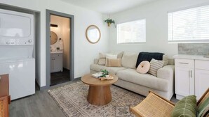 A modern living room with a beige sofa, a round wooden coffee table, and a kitchen area visible through an open door.