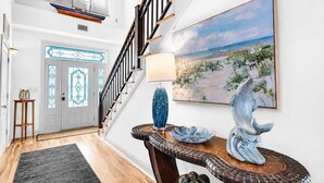 A staircase leading to a second floor, a door with stained glass, a painting of a beach scene, and a decorative table with a blue vase and a ceramic sculpture. - Blessing on the Bay- North Cape Bay Front (Port St. Joe)