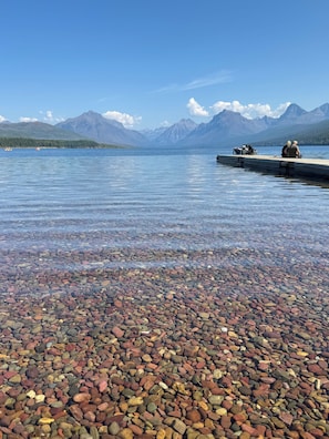 A clear lake with a rocky shore, boats, and mountains in the background.