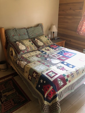 Una cama con colcha que muestra nevados y estrellas, una lámpara y una pared de madera.