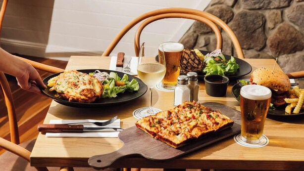 Ein Esstisch mit verschiedenen Gerichten, darunter Pizza, ein Burger und ein Salat, sowie Biergläsern und einer Weinflasche.