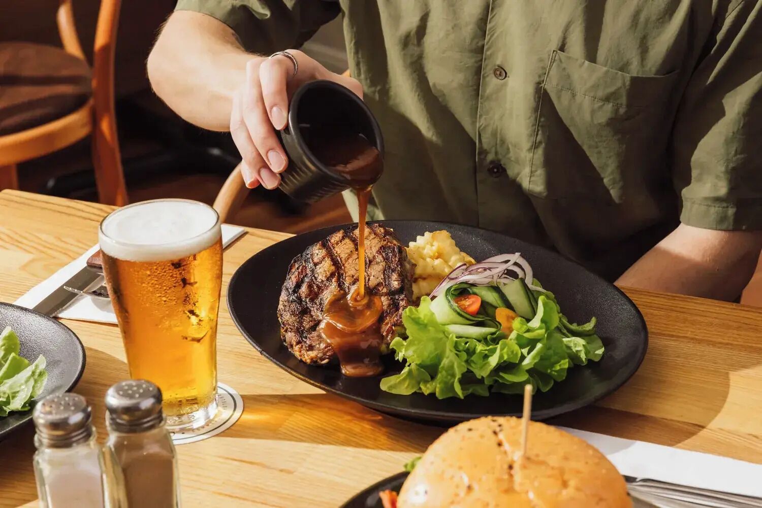 A person pouring sauce over a burger, with a glass of beer and a plate of salad on the table.