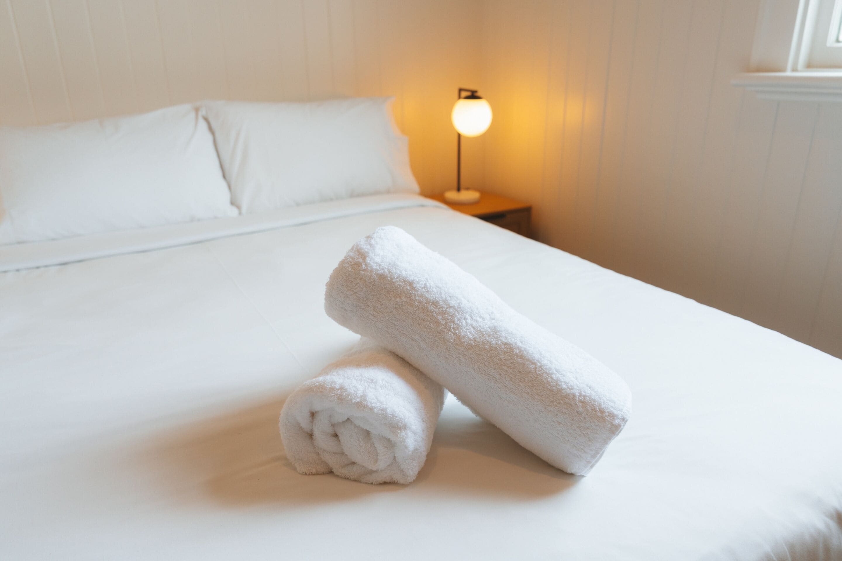 A neatly made bed with white linens and two rolled towels, a bedside lamp, and a window with blinds.