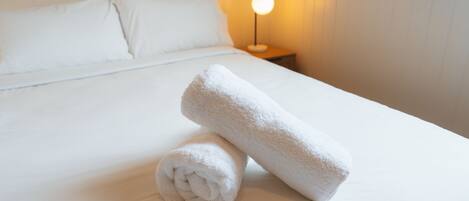 A neatly made bed with white linens and two rolled towels, a bedside lamp, and a window with blinds.