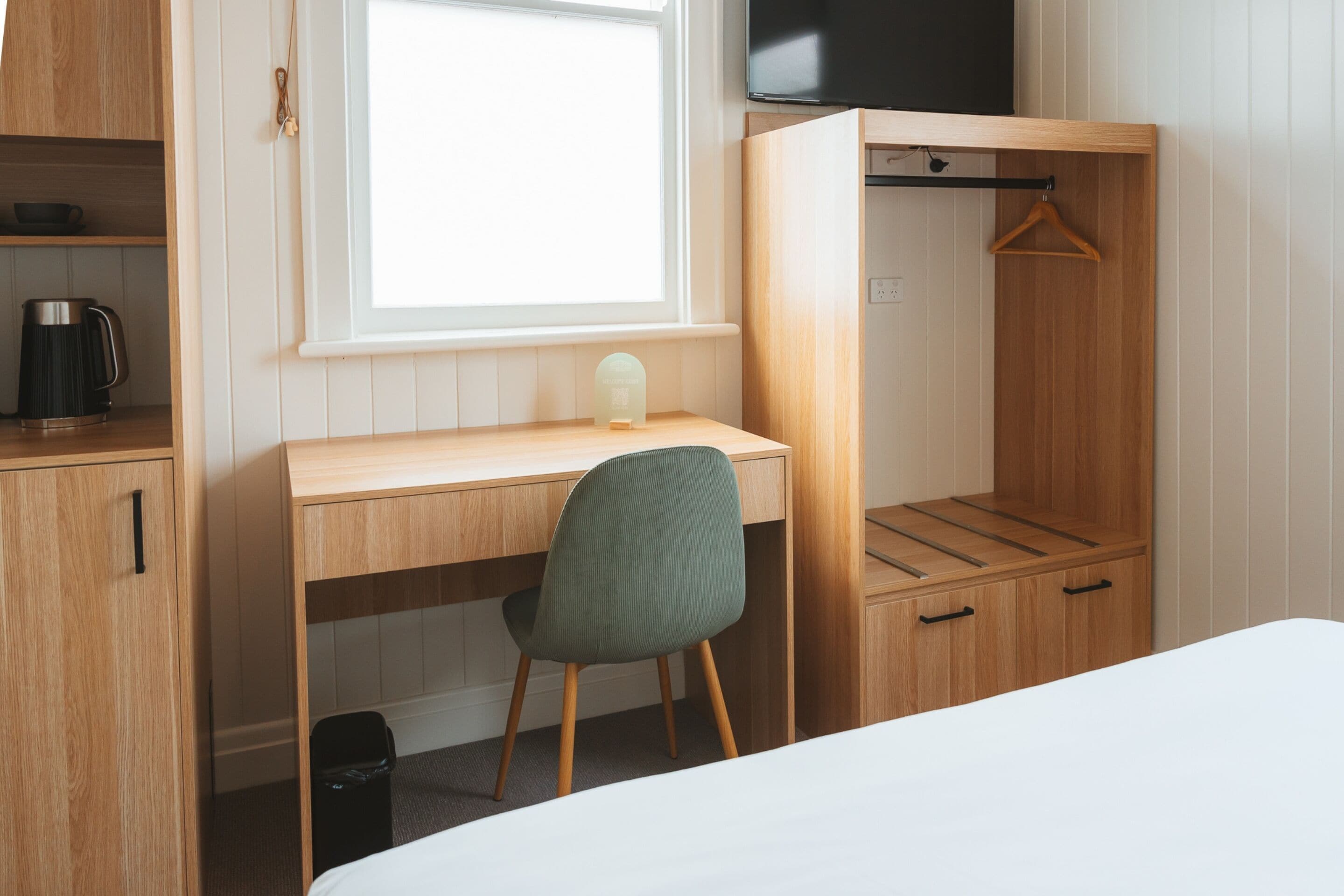 Une pièce comprenant un bureau en bois, une chaise, un lit, un téléviseur et un porte-manteau mural.