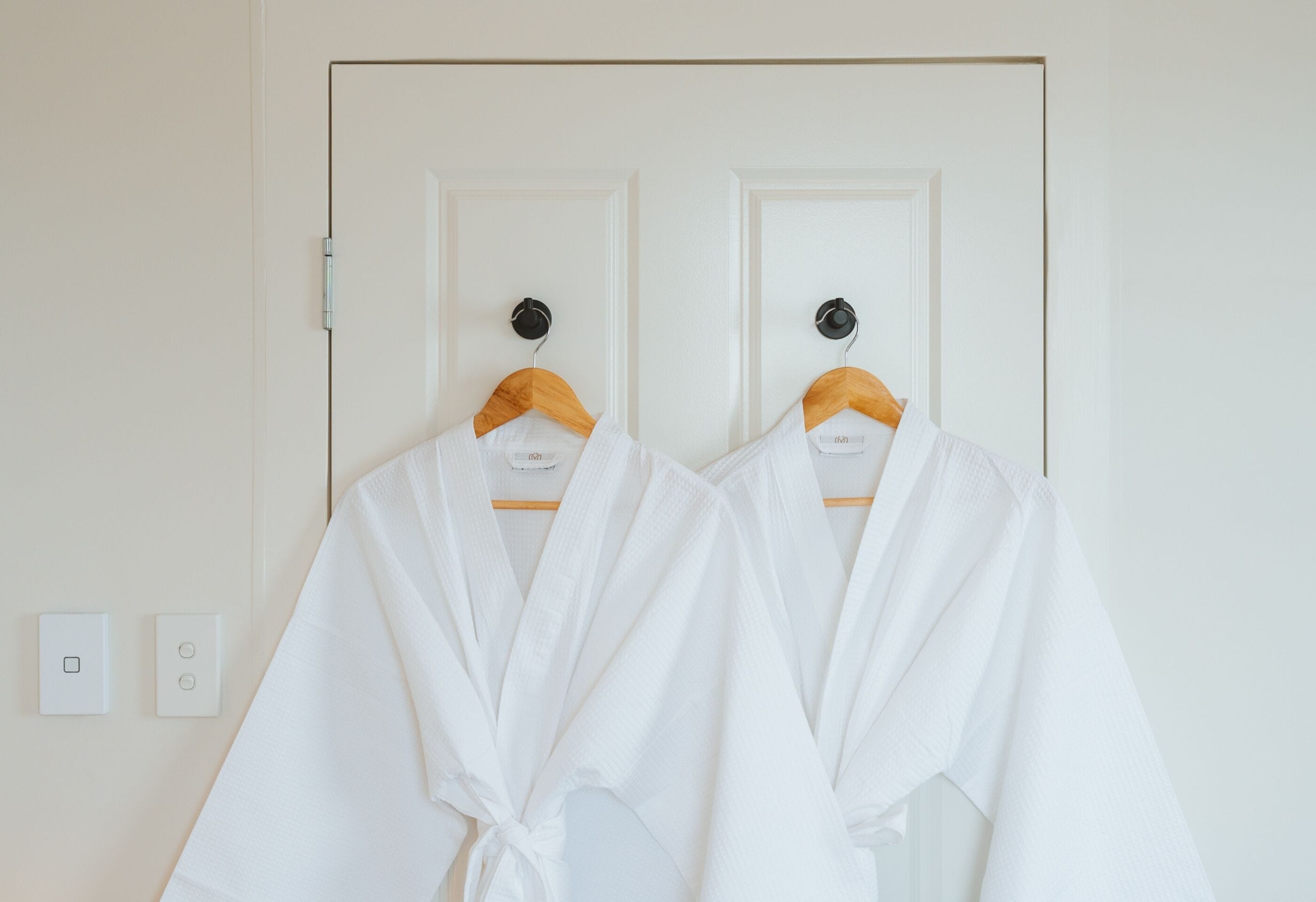 Two white robes hanging on wooden hangers against a white door with a simple light switch and a power outlet visible on the wall.