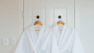Two white robes hanging on wooden hangers against a white door with a simple light switch and a power outlet visible on the wall.