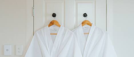 Two white robes hanging on wooden hangers against a white door with a simple light switch and a power outlet visible on the wall.