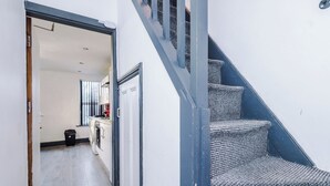 Uma escadaria com carpete texturizado que leva a um patamar. É visível uma cozinha ao fundo, com uma janela e uma porta.