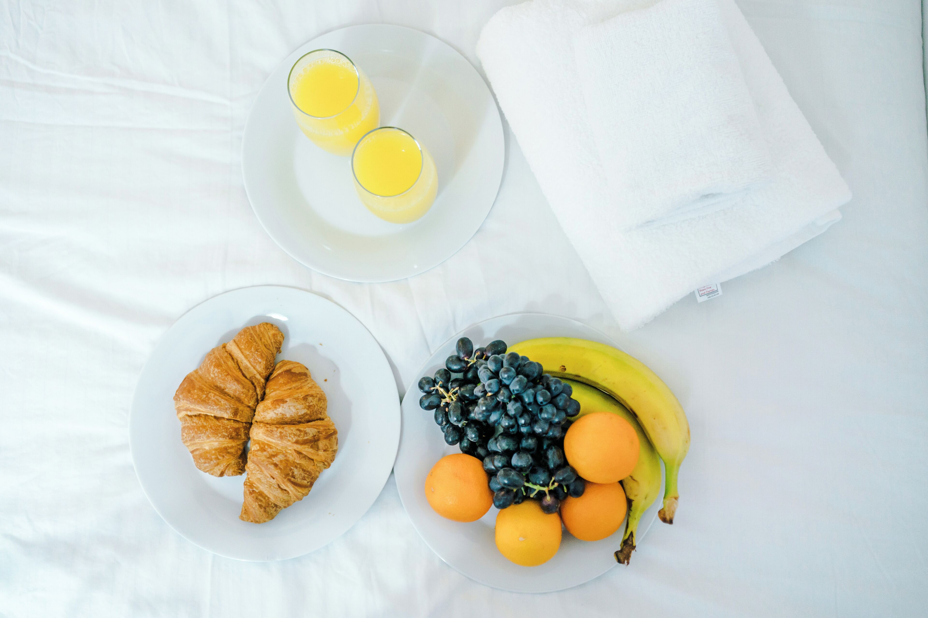 A breakfast setting with croissants, juice, grapes, oranges, and bananas on plates.