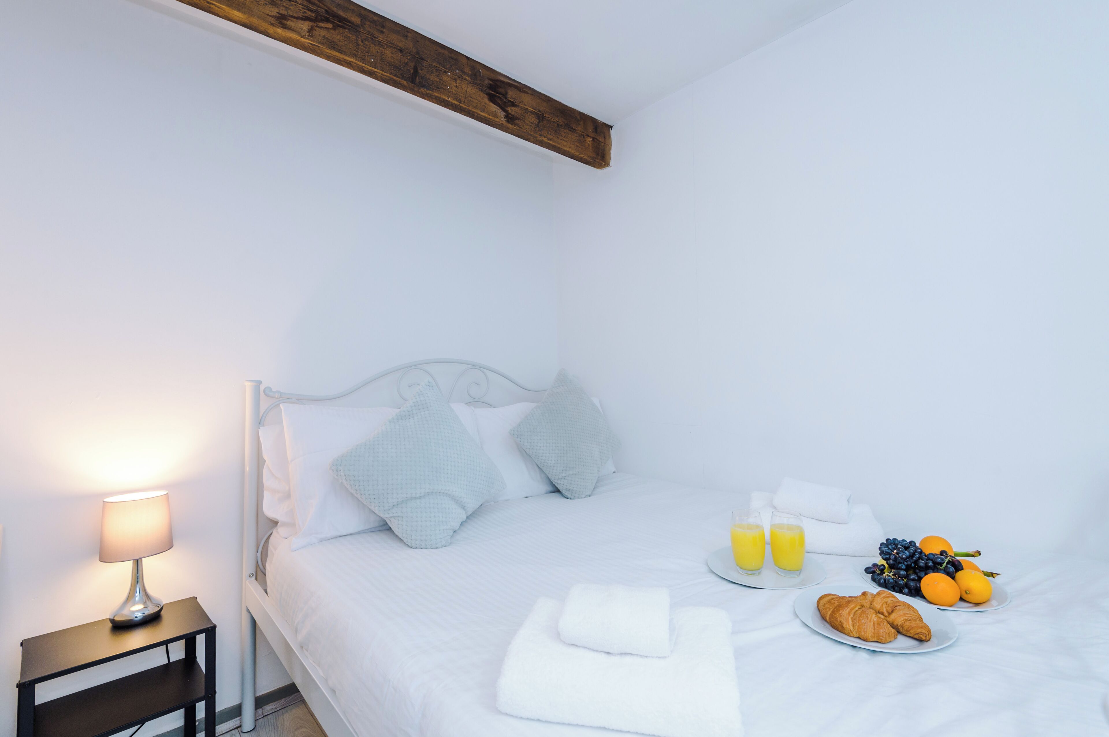 A neatly made bed with pillows, a bedside table with a lamp, and a breakfast tray with croissants, fruit, and juice.