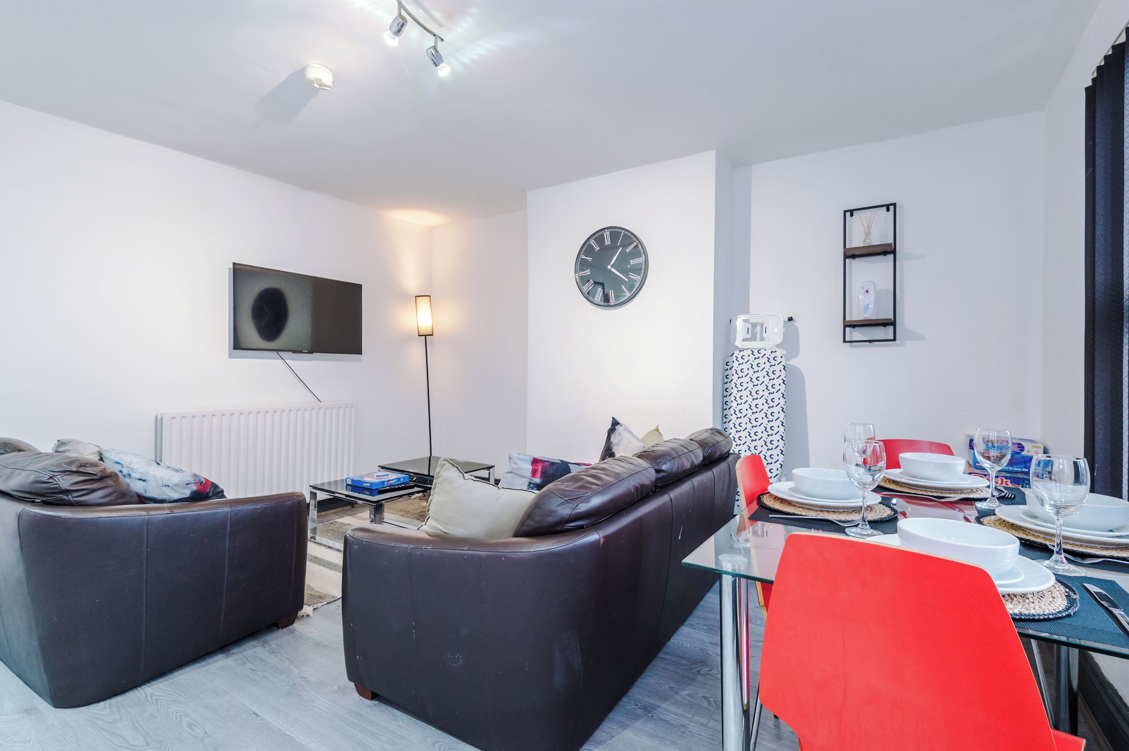 A modern living room with a sofa, a dining table set for two, a flat-screen TV, and a wall clock.
