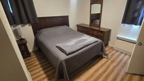 Un lit bien fait, recouvert d’une couverture grise, une commode en bois avec un miroir et une fenêtre avec des rideaux.