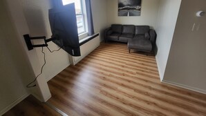 Une pièce avec un plancher en bois, un canapé, un téléviseur fixé au mur et une fenêtre avec des rideaux.