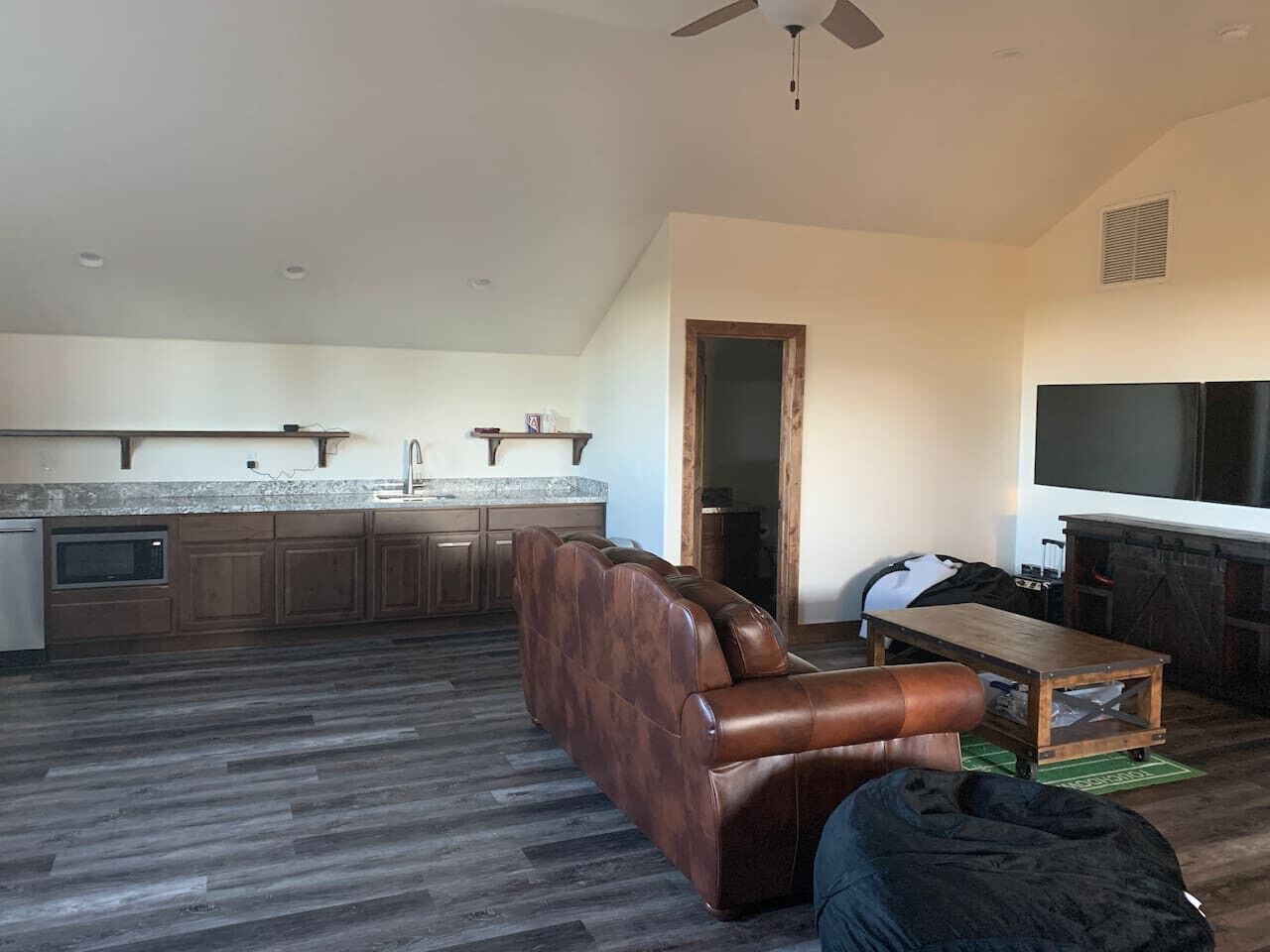 A spacious living area with a brown leather sofa, a wooden coffee table, and a fireplace. A kitchen with wooden cabinets and a microwave is visible in the background.