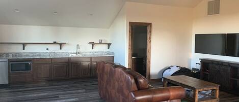 A spacious living area with a brown leather sofa, a wooden coffee table, and a fireplace. A kitchen with wooden cabinets and a microwave is visible in the background.