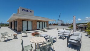 Een terras op het dak met buitenmeubilair, een bordje met de tekst 'PLACON' en een uitzicht op de stad en de haven op de achtergrond.