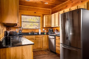 A modern kitchen with wooden cabinets, a stainless steel refrigerator, and a black countertop. - Luxe 5* Retreat~SeptDeals~GmRm~PS5~Views~HotTub (Gatlinburg)