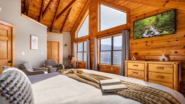 Une chambre spacieuse avec un grand lit, des poutres apparentes en bois au plafond, un téléviseur fixé au mur et de grandes fenêtres offrant une vue sur l’extérieur.