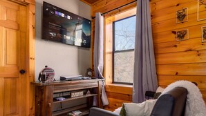 L’intérieur d’un chalet chaleureux, avec un mur en bois, un téléviseur fixé au mur diffusant la série « The Mandalorian », un meuble de rangement en bois et une fenêtre avec des rideaux.