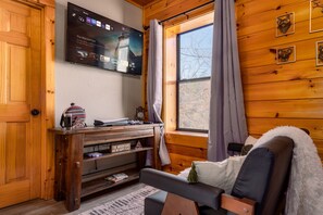 A cozy cabin interior with a wooden wall, a mounted TV displaying 'The Mandalorian', a wooden entertainment unit, and a window with curtains. - Luxe 5* Retreat~SeptDeals~GmRm~PS5~Views~HotTub (Gatlinburg)